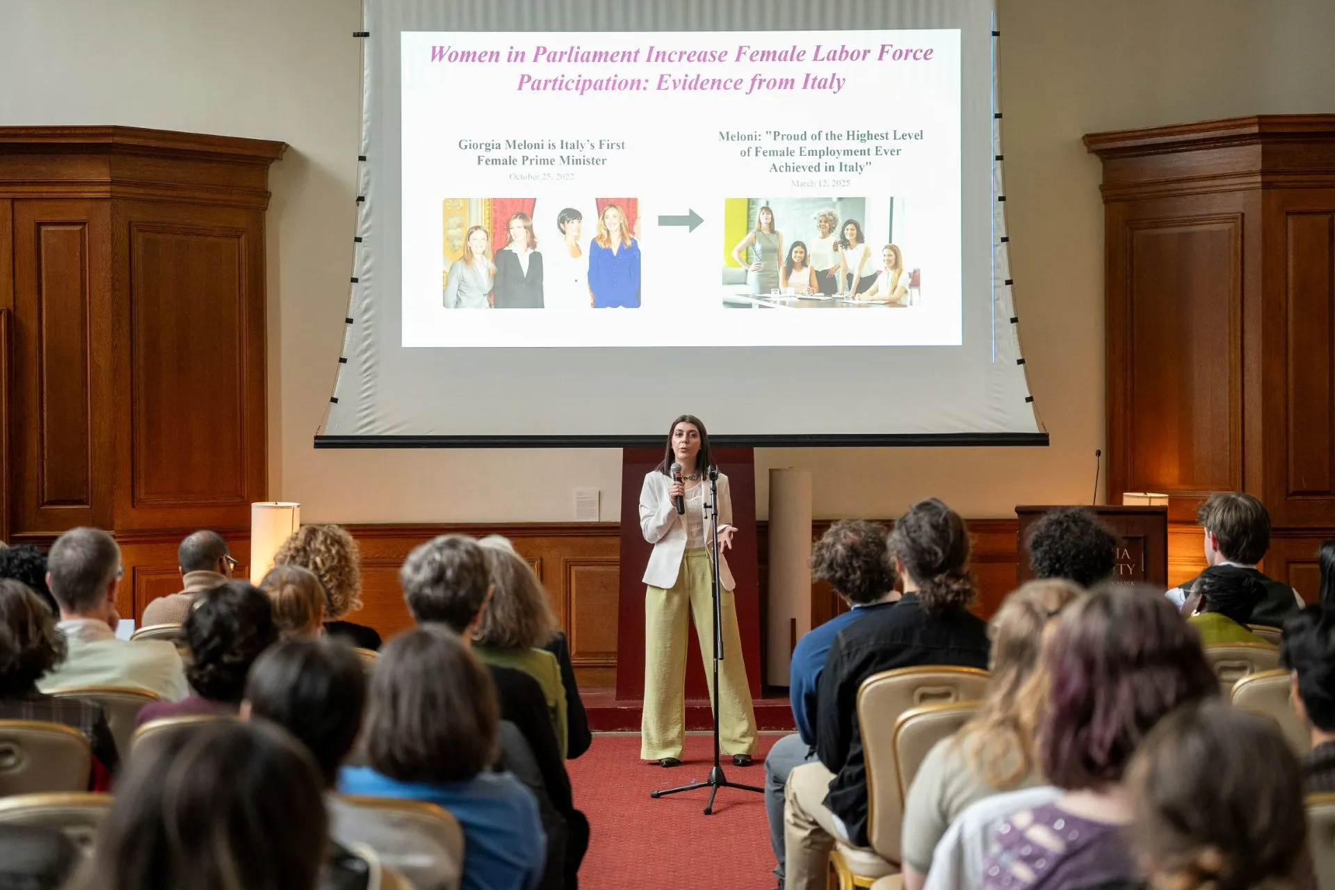 A woman in a pantsuit speaks into a microphone with a slideshow behind her of women in parliament in Italy. 