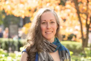 Head and shoulders of woman with long brown curly hair, wearing a scarf and smiling to camera. Campus greenery in background. 