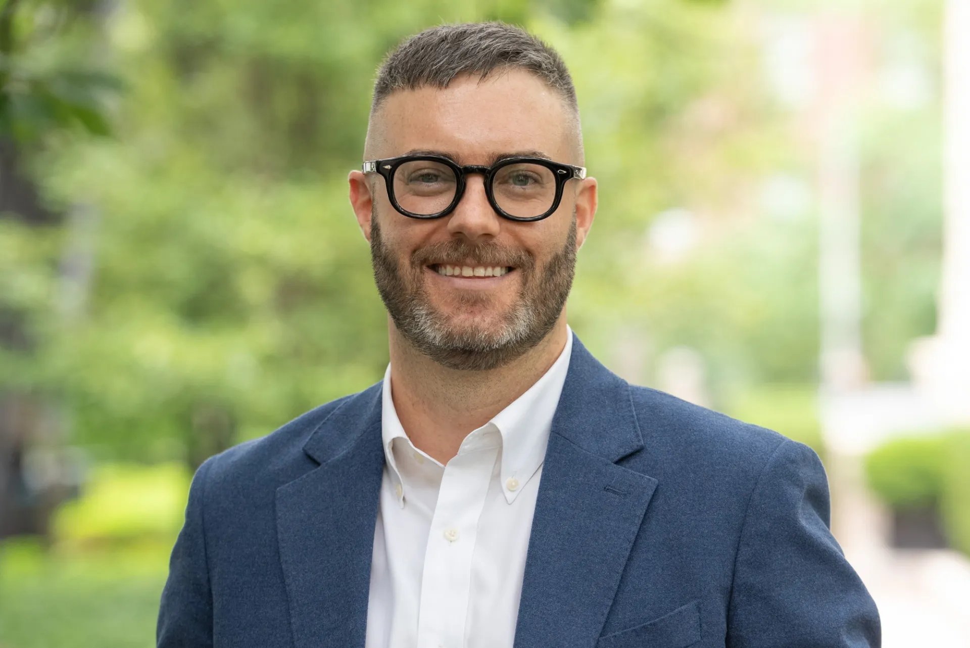 A professional headshot of a man with short light brown hair and a beard, with dark glasses, wearing a blazer and button-up, taken outdoors.