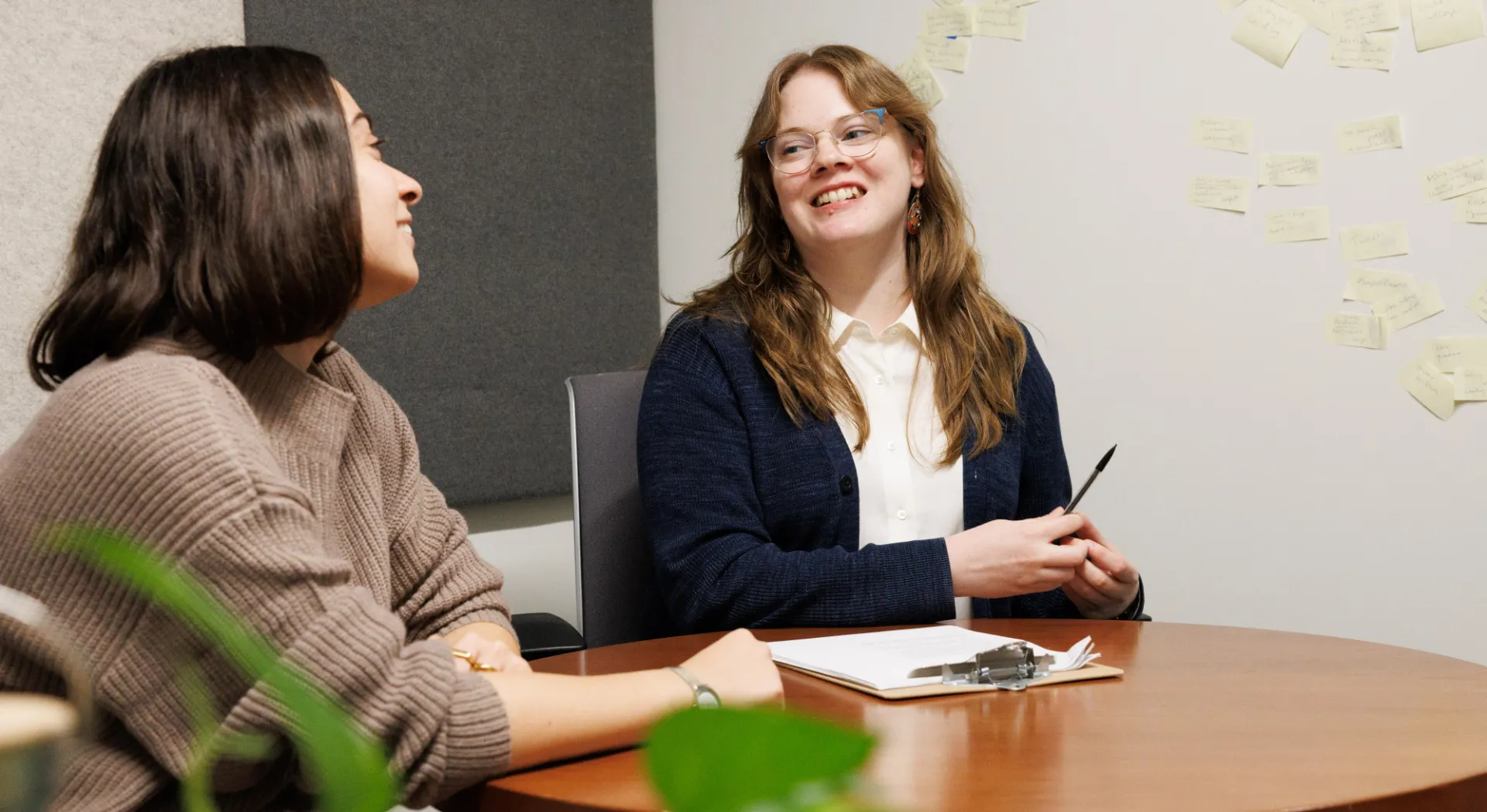 Two women sit at a desk with a pen and paper in front of them, smiling at one another. There is a wall with post-its stuck to it behind them, and a leafy plant in the foreground. The mood is studios and lighthearted.
