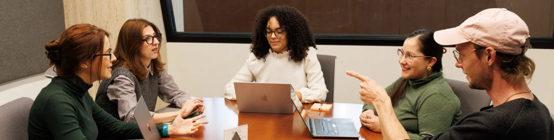 Five students gather at a conference table, with a window behind them that lets in bright light. They have laptops and notebooks open, as they discuss a question with serious and smiling faces.